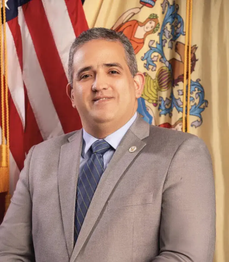 Official portrait of Carlos Rivas in front of the United States and New Jersey flags